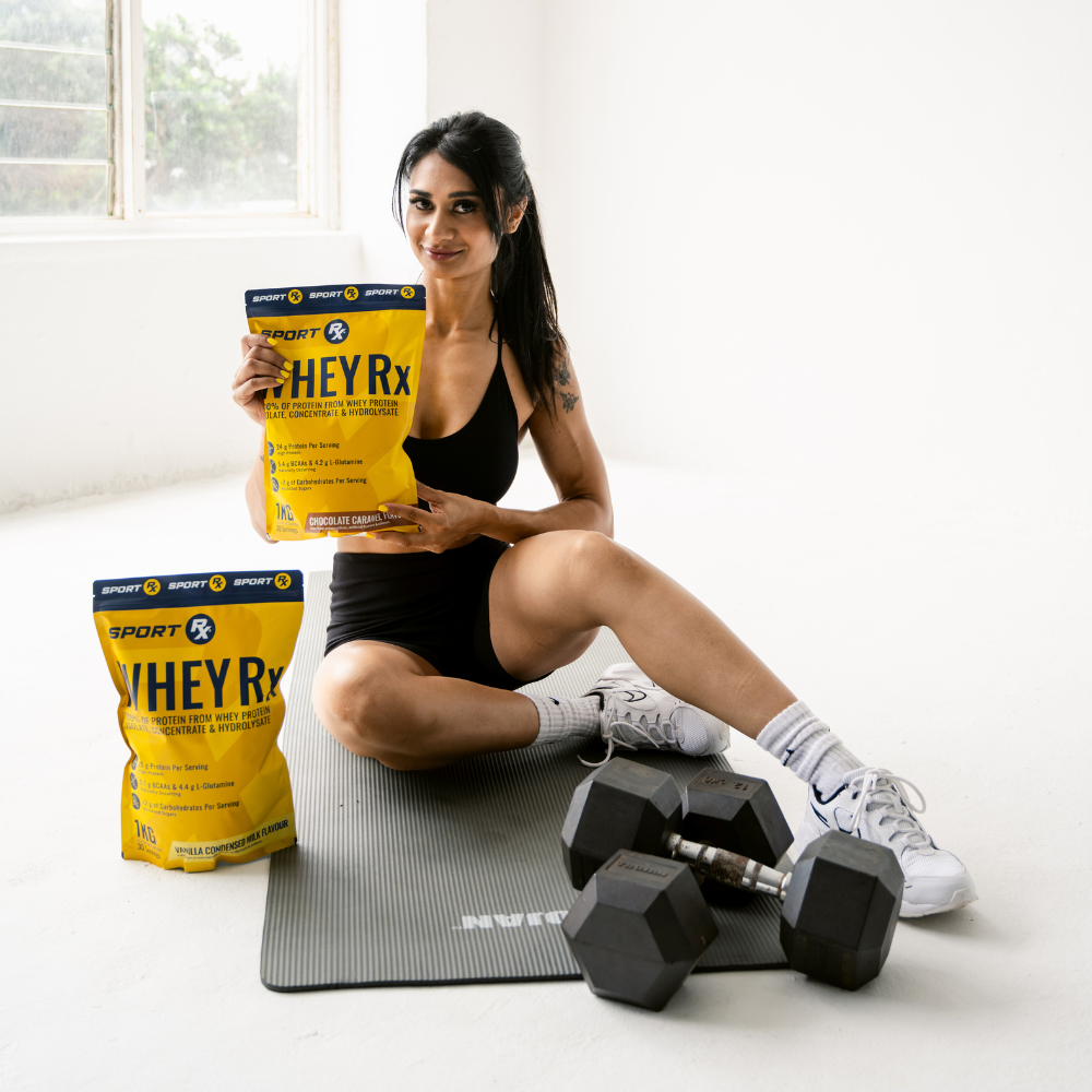 Woman holding a Sport Rx WHEYRx protein powder bag with dumbbells and a mat on a light background