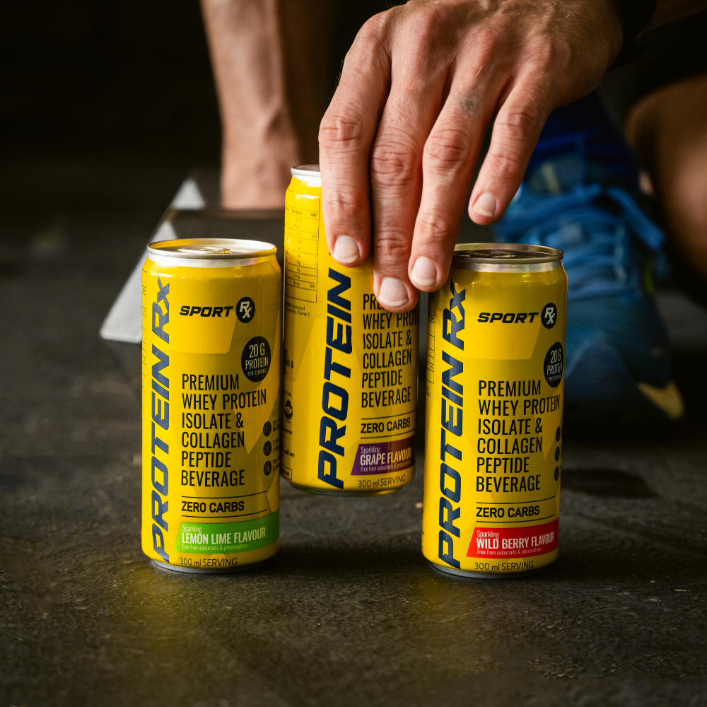 Three cans of Protein X brand protein drinks on a dark surface with a person's hand reaching towards them.